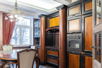 wooden kitchen unit in brown color. Built-in large kitchen with upper and lower cabinets. Large wooden dining table and chairs. Metal chandelier, dark wallpaper, wooden doors.