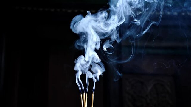 Mystical aroma of burning incense sticks releasing swirling wisps of smoke against a dark backdrop creating abstract shapes and patterns