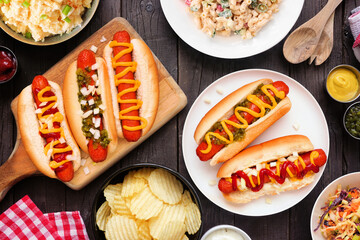 BBQ table scene with hot dogs, classic salads and snacks. Above view over a dark wood background.