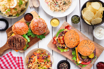BBQ table scene with hamburgers, classic salads and snacks. Top down view over a white wood background.