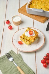 Sliced ​​cabbage, carrot and egg casserole on a plate on a light wooden background. Served with sour cream and fresh cherry tomatoes.