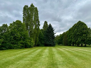 A wide, freshly mowed lawn stretches forward with perfectly aligned stripes, leading the eye toward a variety of tall, lush trees in the background. The cloudy sky above adds a dramatic contrast to th