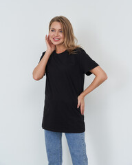 Smiling woman with blonde hair in casual black t-shirt and blue jeans, one hand on hip and other touching her face, looking to the side. Light gray background