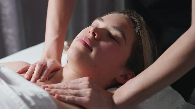 Massage therapist in black uniform gently massaging collarbone and upper chest of light-skinned female client lying with eyes closed on spa bed, showing peaceful relaxation under soft natural light