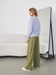 A woman with light brown hair in a blue sweater and olive-green pants stands sideways. She looks at a full-length mirror, while a stylish sofa is visible in the background