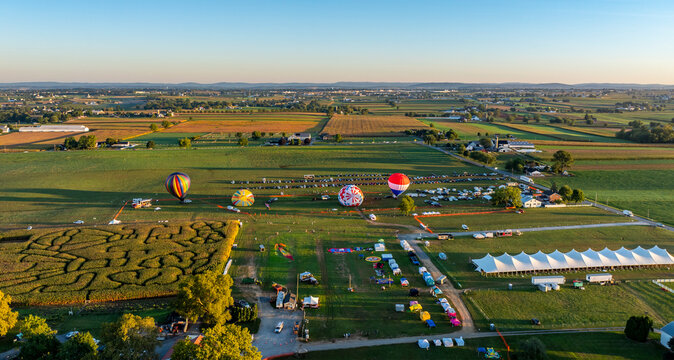Hot air balloons take flight above expansive fields during a vibrant festival. The sky is painted with hues of orange as vendors and visitors enjoy various activities beneath.