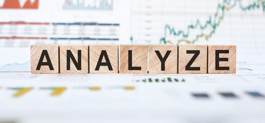 Wooden blocks spelling the word analyze on a white table with blurred financial charts in the background.