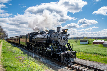 Naklejka premium A powerful steam engine makes its way along a rural track, releasing clouds of steam into the bright blue sky. Fields stretch out on either side, dotted with white barns.