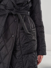 Quilted black coat with a belted waist and large front pocket, worn by an individual. The design...