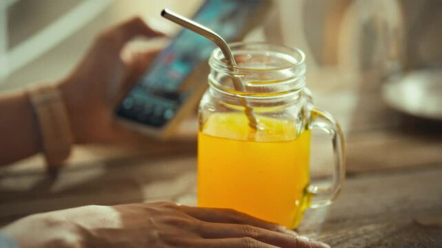 A hand rests on a wooden table, holding a glass mug filled with a bright yellow drink. Nearby, a smartphone shows a lively screen, capturing a relaxed afternoon atmosphere