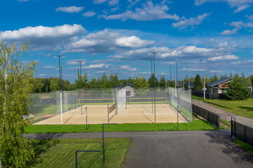 Obraz premium A spacious outdoor sports court with sandy flooring is surrounded by a mesh fence, featuring bright lighting poles. Lush green trees and houses are seen in the background under a sunny sky.
