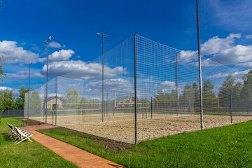 A spacious volleyball court features sandy ground and tall nets, set amidst vibrant green grass and a clear blue sky. A wooden pathway leads to the court, enhancing the recreational atmosphere.