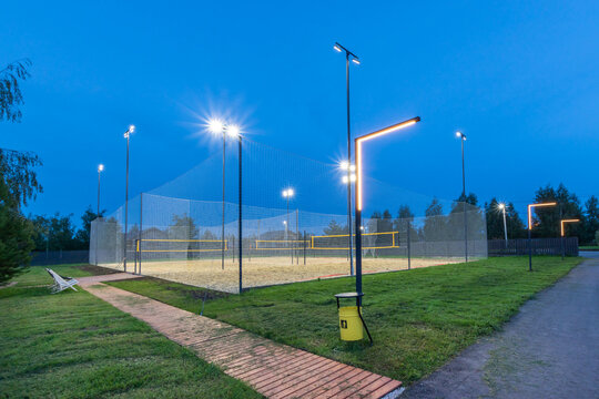Bright lights illuminate a sand volleyball court during dusk. The area is surrounded by greenery and features a well-maintained pathway leading to the court.