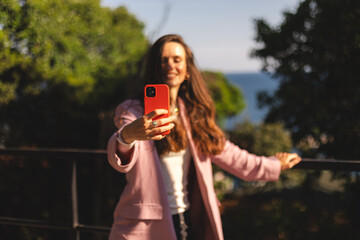 Attractive long hair brunette woman walking outdoor against sea landscape, make video call, or selfie photo, enjoy. Girl wear pink blazer. Focus on phone.