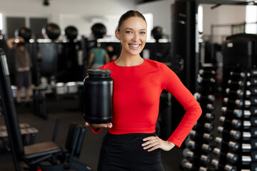 Fototapeta premium Attractive woman holding big protein jar after workout posing and smiling in modern fitness gym interior, ad for protein and additional fitness nutrition