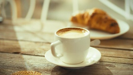 A cup of coffee sits on a white saucer beside a freshly baked croissant. Sunlight streams through a window, casting warm light on the wooden table in the cozy cafe
