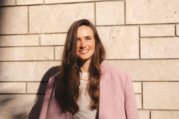 Woman entrepreneur with crossed hands standing on office building background. Successful smiling caucasian woman in business suit, pink blazer on shoulders. Female worker with crossed arms look happy.
