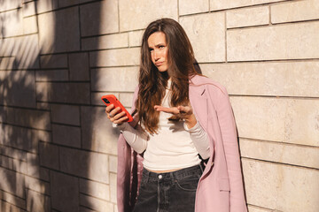 Mad and shock brunette beautiful woman using mobile phone standing near wall office building. Yelling unhappy woman hold mobile phone. Disappointed sad upset lady horrified impressed news. WTF. Oh no