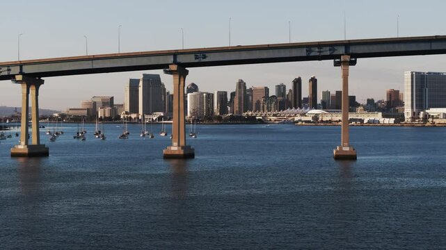 Aerial: Drone Panning Shot Of Cars Moving On Famous Coronado Bridge By Modern Buildings In City Against Clear Sky - San Diego, California