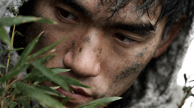 Closeup portrait of a mans face covered in dirt and rain partially obscured by foliage