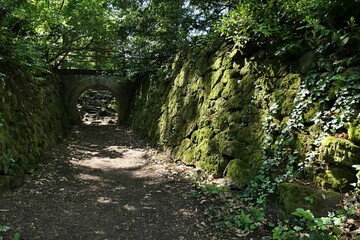Park old stone arc with pathway and railing above another path with moss covered stone walls around. Late spring daylight sunshine and shadows from surrounding foliage visible.  