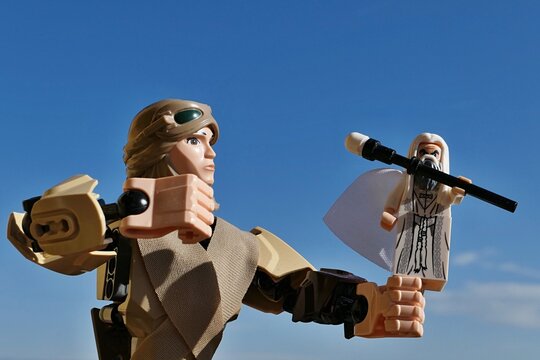 LEGO Star Wars action figure of female jedi Rey Skywalker in scavenger outfit aiming to punch small figure of LEGO Saruman from Lord Of The Rings movie with her right fist. Blue skies background. 