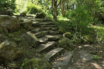 Fototapeta premium Old partially damaged stone stairs with mossy rocks around, hidden in shadow of park trees, spring daylight sunshine. 