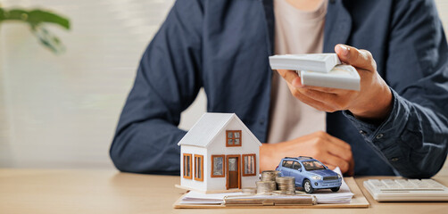 Money bills held above house and car models with paperwork. Represents tax-saving strategies, cash...