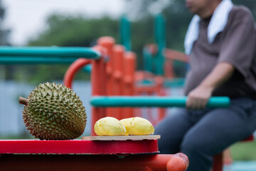 Fototapeta premium A whole and peeled durian sit on outdoor gym equipment as a man exercises in the background. The image connects health, fitness, and dietary choices, highlighting durian’s nutritional impact.