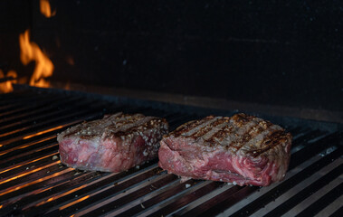 Close-up of two pieces of meat sizzling on the grill over an open flame. The fire crackles beneath the juicy steaks, capturing the essence of outdoor cooking.