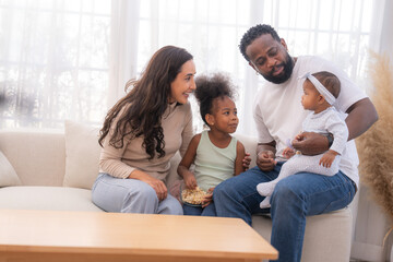 Fototapeta premium Happy family sitting together on a sofa, spending quality time and smiling while enjoying snacks at home in a warm and cozy living room.