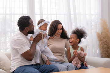 Fototapeta premium Happy mixed race family sitting on the living room floor, smiling and enjoying time together at home with their kids and baby in a warm, bright, cozy atmosphere.