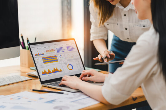 Two women working together on data analysis using laptop with colorful charts and graphs on screen, discussing business strategy in modern office environment