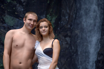 A young semi-naked couple looks at the camera, standing by a waterfall in a grotto. Close-up. Copy space
