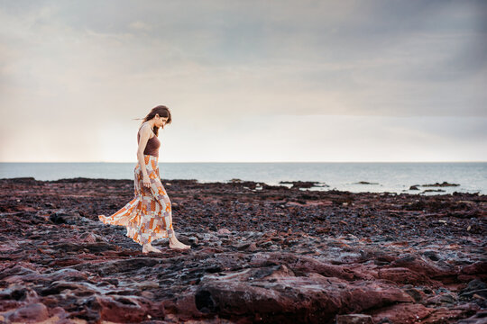 A woman wearing a long skirt walking on a rocky shore with ocean as the background