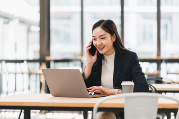 Fototapeta premium Young Asian businesswoman smiling while talking on smartphone and working on laptop in modern office, enjoying productive remote work with coffee