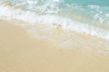 A close-up shot of a sandy beach meeting the clear turquoise ocean. Gentle waves are breaking on the shore, creating a peaceful and inviting atmosphere. The image captures the beauty of the coast