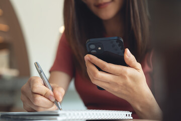 Woman in casual business clothes using smartphone, writing on notebook at coffee shop, digital nomad. Student using mobile phone, online studying via laptop, e-learning, remote work from cafe