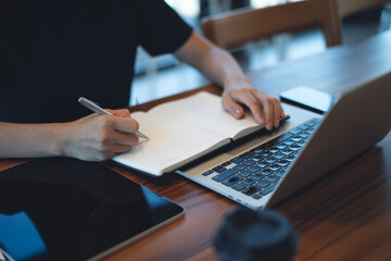 Close up of business woman hand with a pen writing, planning on notebook, working on laptop computer on table at coffee shop. Student studying online, taking note on notepad, e-learning