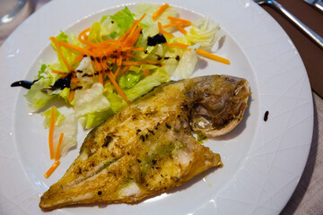 Halved fried dorada fish served on white plate with vegetable salad and balsamic sauce
