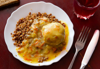 Boiled buckwheat with meatball and sauce served in white plate with serving pieces.