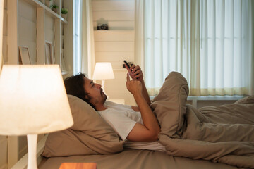 Fototapeta na wymiar Asian man in white shirt lays in bed using and looking at mobile phone as soft morning light filters through the curtains. People with insomnia.
