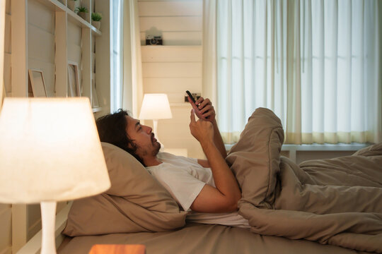 Asian man in white shirt lays in bed using and looking at mobile phone as soft morning light filters through the curtains. People with insomnia.
