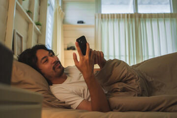 Bearded Asian Japanese man lying comfortably in bed, smiling while using smartphone and enjoying a...