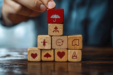 Hand stacking wooden blocks with various red icons including hearts, arrows, a clock, a caution sign, a person symbol, and an umbrella symbol on top, representing protection and care