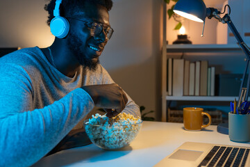 Happy black man watching movie, eating popcorn at night wearing headphones.