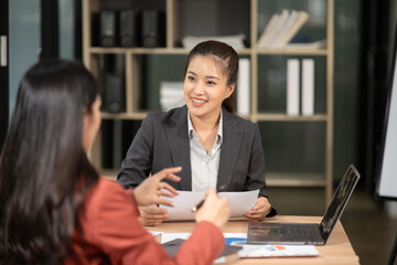 Two Asian businesswomen with laptops discussing documents in an office.
