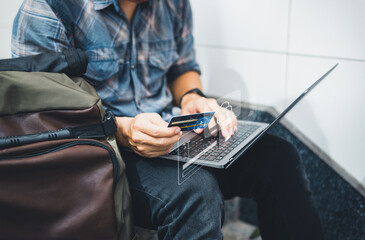 A man sitting on the floor with a backpack, using a laptop and credit card for online payment,...