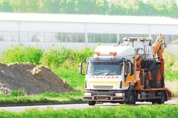 Orange tow truck hauling mini excavator on a rural road in summer