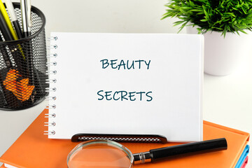 A notebook showcasing the words beauty secrets sits on a stack of colored books in a tidy workspace. A magnifying glass and potted plant add charm to this inspiring atmosphere for beauty enthusiasts